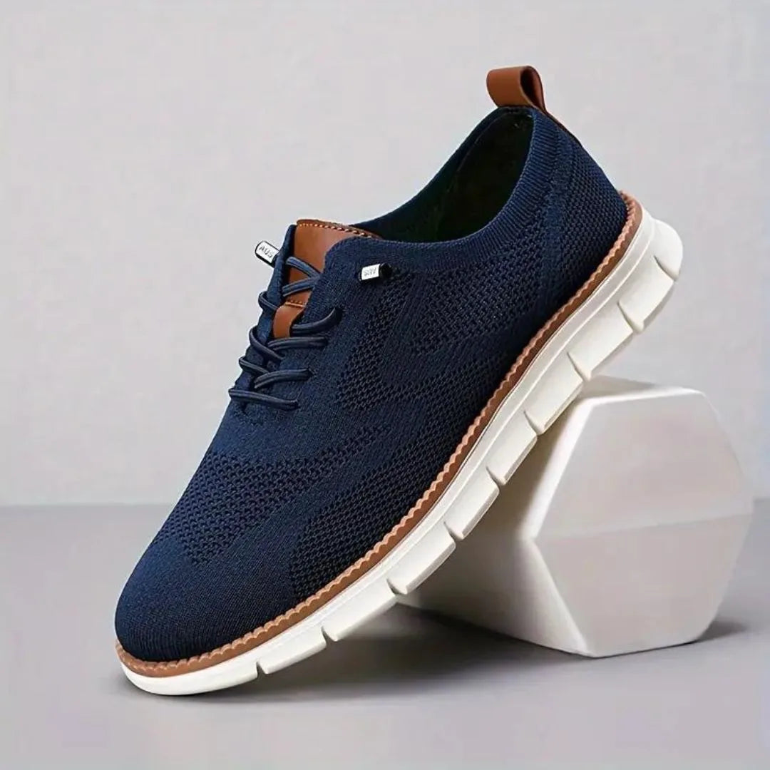 Navy blue knit sneaker with tan heel loop and lace eyelets resting against a white geometric block
