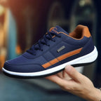 Hand holding a navy blue leather trainer with tan panels and white chunky sole, showing side logo badge