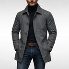 Man wearing a grey mac-style trench coat open over a black polo neck jumper and jeans