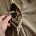 Close-up of a hand opening the welt pocket on a beige trench coat to show a black horn-style button