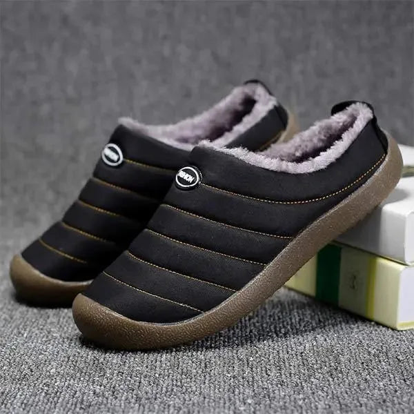 Side view of a black quilted slip-on slipper with grey faux fur trim and brown sole