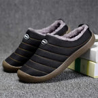 Side view of a black quilted slip-on slipper with grey faux fur trim and brown sole