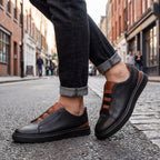 Side profile of a man walking on a cobbled street wearing black leather slip-on trainers with tan heel trim