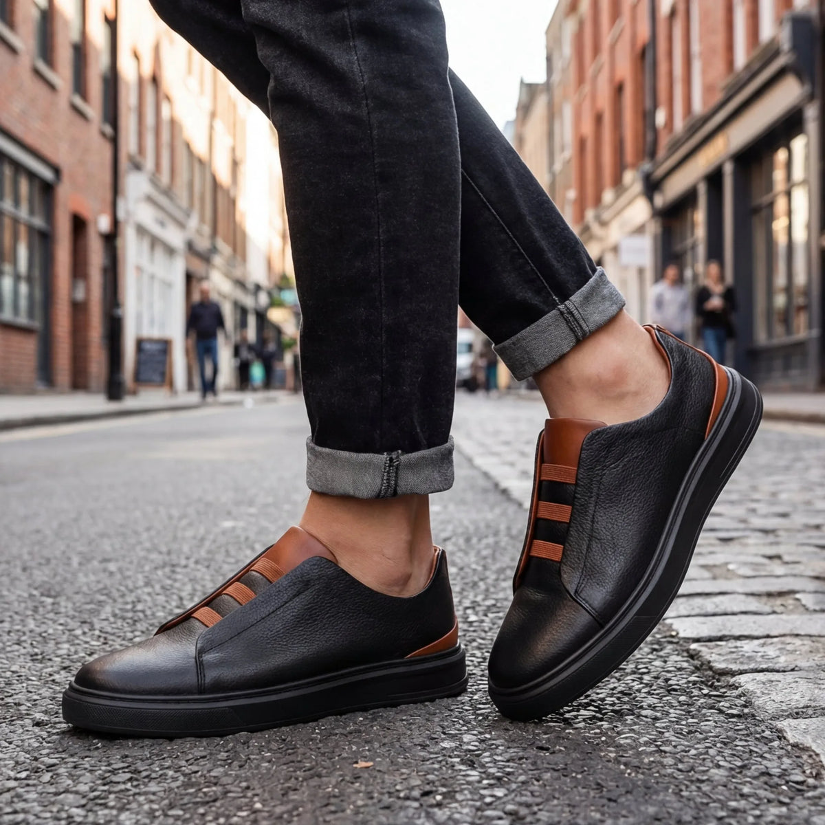 Side profile of a man walking on a cobbled street wearing black leather slip-on trainers with tan heel trim