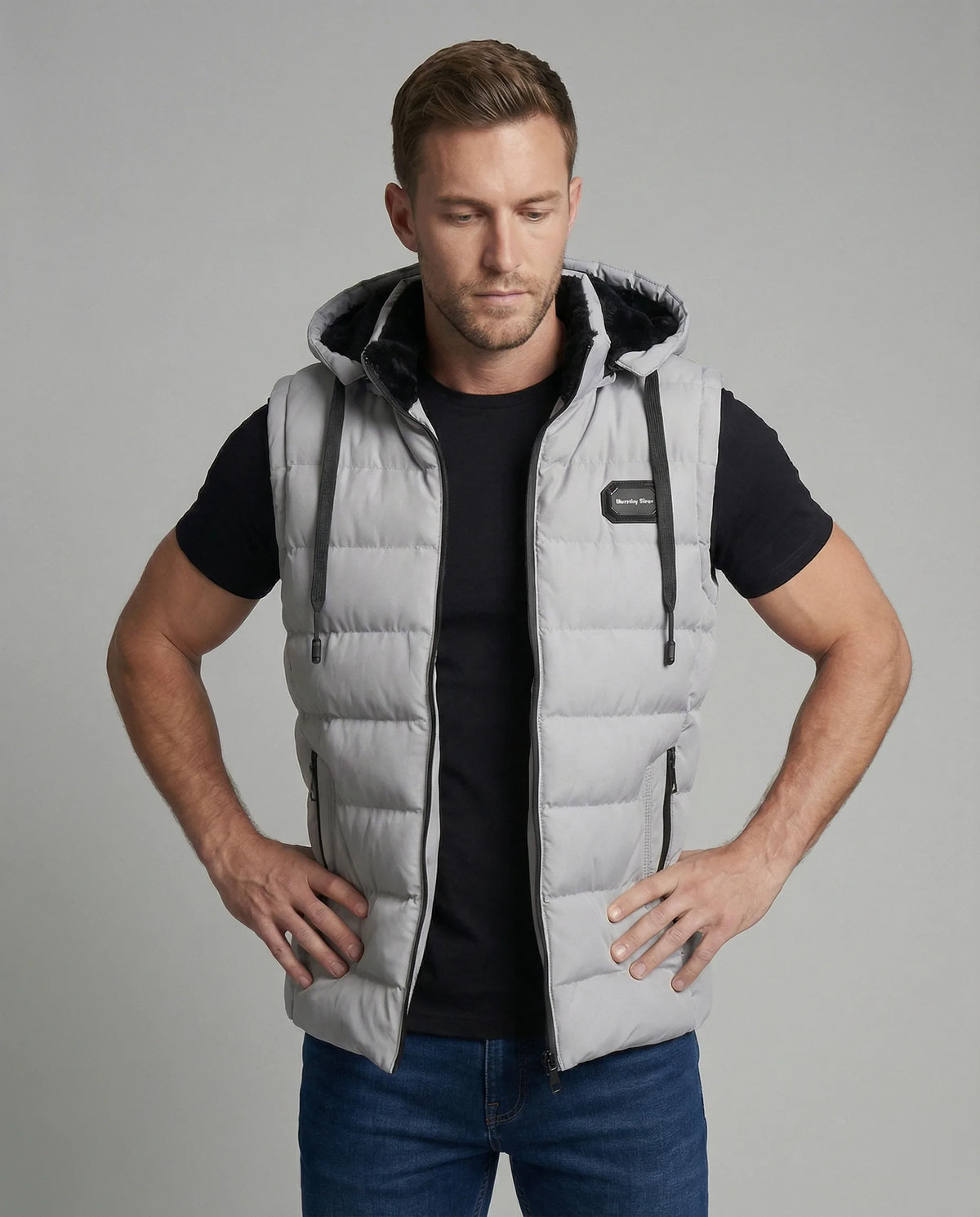 Man wearing a grey hooded quilted gilet with faux fur hood lining over a black t-shirt and jeans, front view