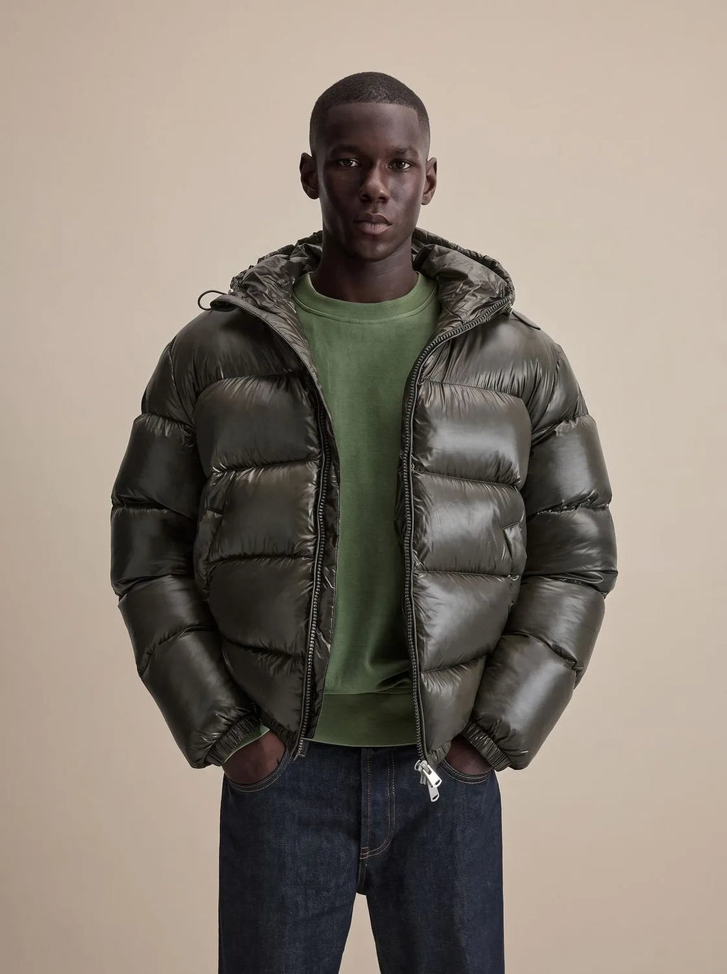 Man wearing a glossy dark olive hooded puffer jacket, unzipped over a green jumper, hands in pockets