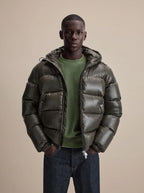 Man wearing a glossy dark olive hooded puffer jacket, unzipped over a green jumper, hands in pockets
