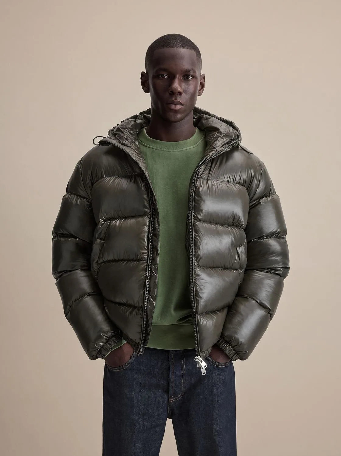 Man wearing a glossy dark olive hooded puffer jacket, unzipped over a green jumper, hands in pockets