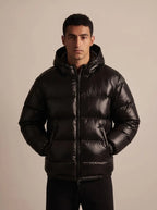 Man modelling the glossy black puffer jacket fully zipped up with the hood resting on his shoulders