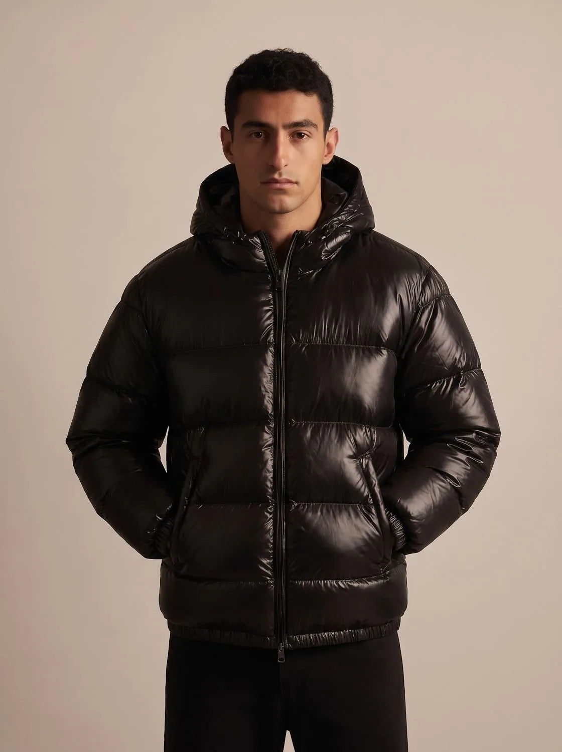 Man modelling the glossy black puffer jacket fully zipped up with the hood resting on his shoulders