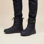 Close-up of feet wearing black insulated snow boots with side zip over dark trousers on a beige background