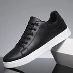 Side view of a black leather lace-up trainer with a padded ankle collar, black laces and a white sole, resting against a grey block.