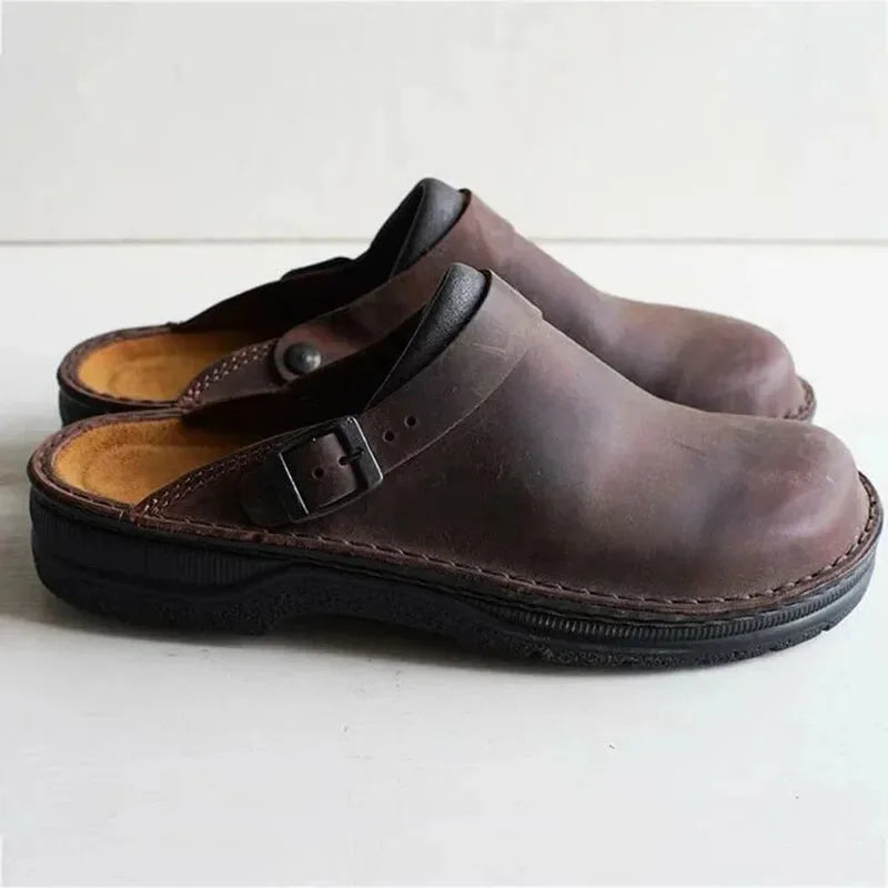 Side view of dark brown leather clog mules with a buckled strap and thick black sole, worn together on a wooden floor