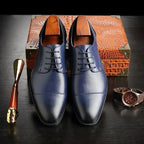 Front view of navy blue leather lace-up dress shoes with cap-toe detail and wooden shoe trees, set against a crocodile-textured case