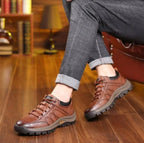 Brown leather shoe worn on foot while seated, paired with grey trousers on a wooden floor