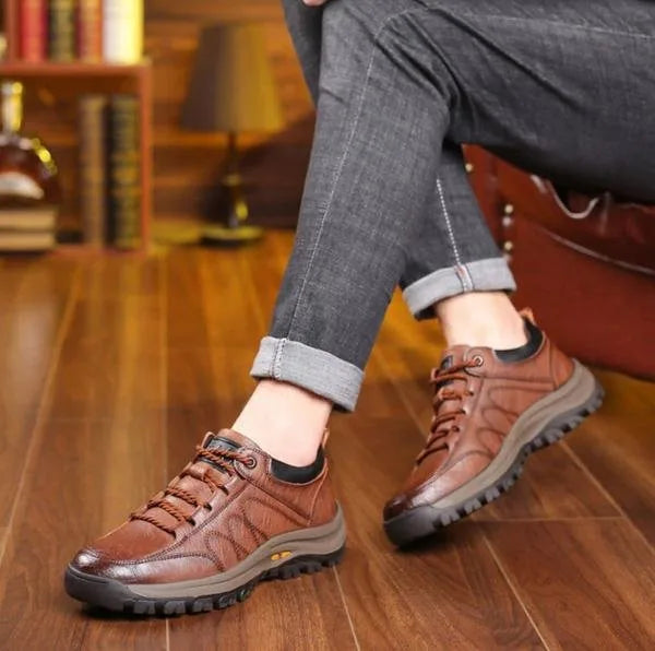 Brown leather shoe worn on foot while seated, paired with grey trousers on a wooden floor
