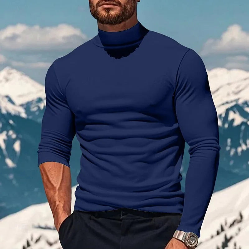 Man in a navy blue mock neck long sleeve top, paired with dark trousers, mountains in the background