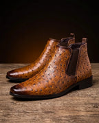 Pair of brown ostrich-embossed leather Chelsea boots on a wooden floor, showing pointed toe and pull-tab heel detail