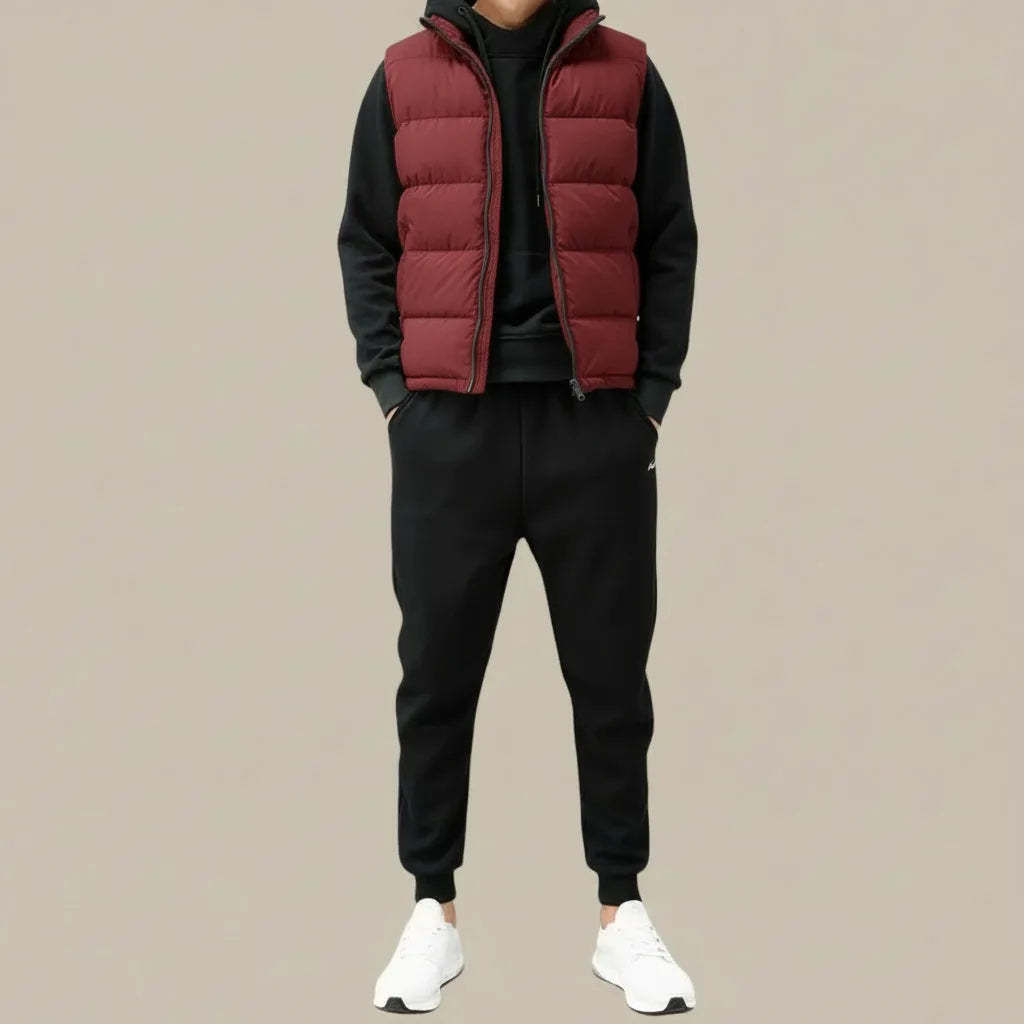 Man wearing a burgundy quilted gilet zipped over a black hoodie, paired with black joggers and white trainers