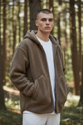 Front view of a man wearing a brown textured sherpa fleece zip hoodie with hood up over a white t-shirt in a forest setting