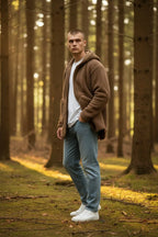 Side angle of the brown sherpa fleece hoodie showing the relaxed fit, front pockets and zip detail against a forest backdrop