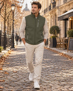 Man walking in a khaki green softshell gilet layered over a cream knit jumper with beige trousers and white trainers.