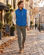 Man strolling in a bright blue softshell gilet layered over a cream jumper with khaki trousers and brown boots.