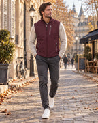 Man in a burgundy softshell gilet with zipped pockets layered over a cream jumper, grey trousers and white trainers.