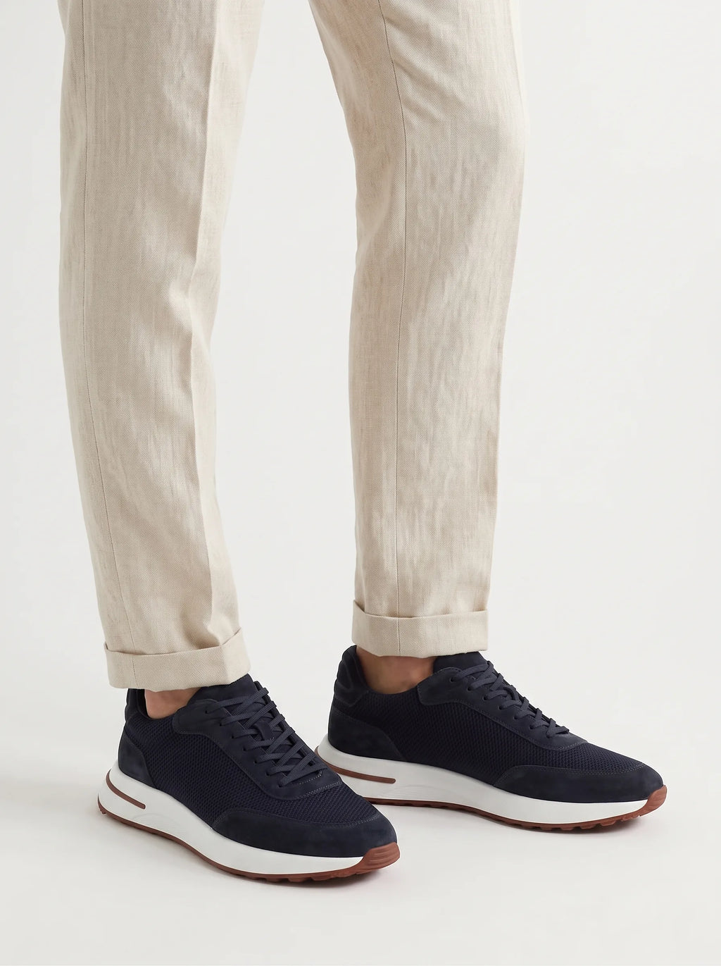 Side view of navy suede trainer with mesh panel, white midsole and brown trim, worn with cream turned-up trousers