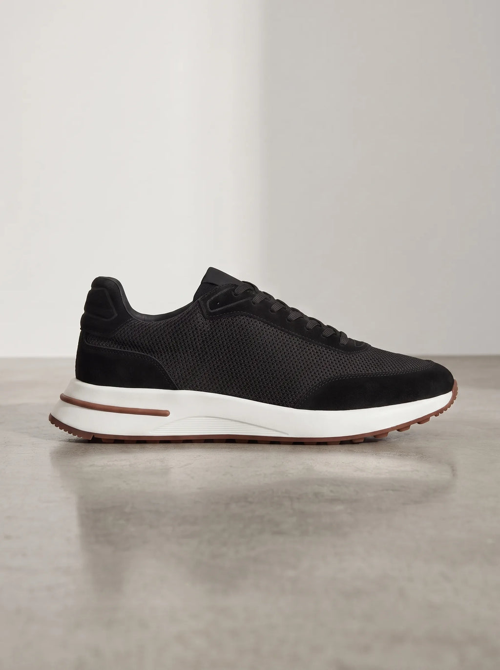 Side profile of a black suede trainer with mesh panel, white midsole and brown stripe detail on a concrete floor