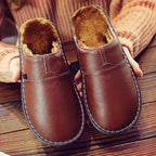 Pair of brown leather mule slippers displaying warm fleece lining and hand-stitched detailing on a wooden floor.
