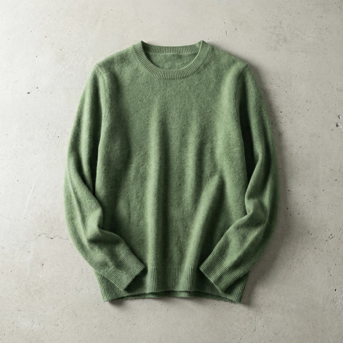 Folded sage green merino wool jumper with ribbed crew neckline, laid flat on a textured grey backdrop