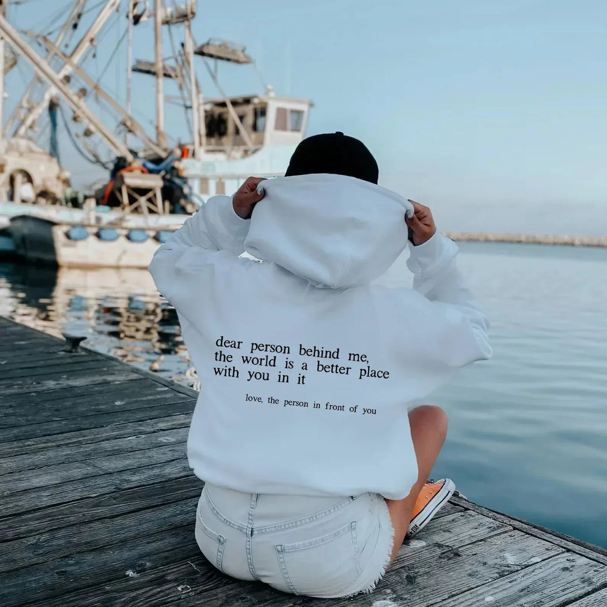 Back view of a white oversized hoodie worn on a wooden pier, showing the lowercase back message in full