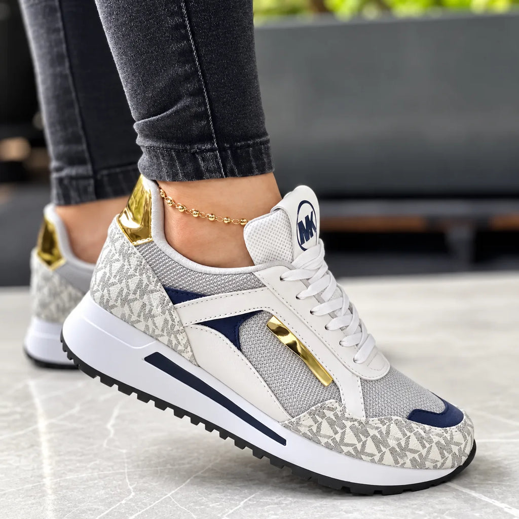 White and grey monogram print trainer with navy suede panel and gold lace trim, worn outdoors on stone flooring