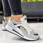 White and grey monogram print trainer with navy suede panel and gold lace trim, worn outdoors on stone flooring
