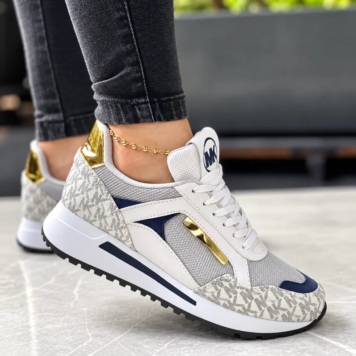 White and grey monogram print trainer with navy suede panel and gold lace trim, worn outdoors on stone flooring