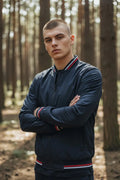 Man wearing a navy bomber jacket with arms crossed, showing striped ribbed collar and cuffs in a forest setting.