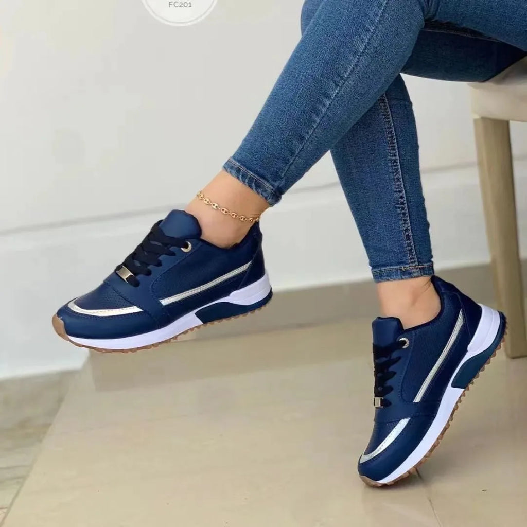 Navy blue trainers with silver side stripe and white chunky sole, worn by a seated model with rolled-up jeans