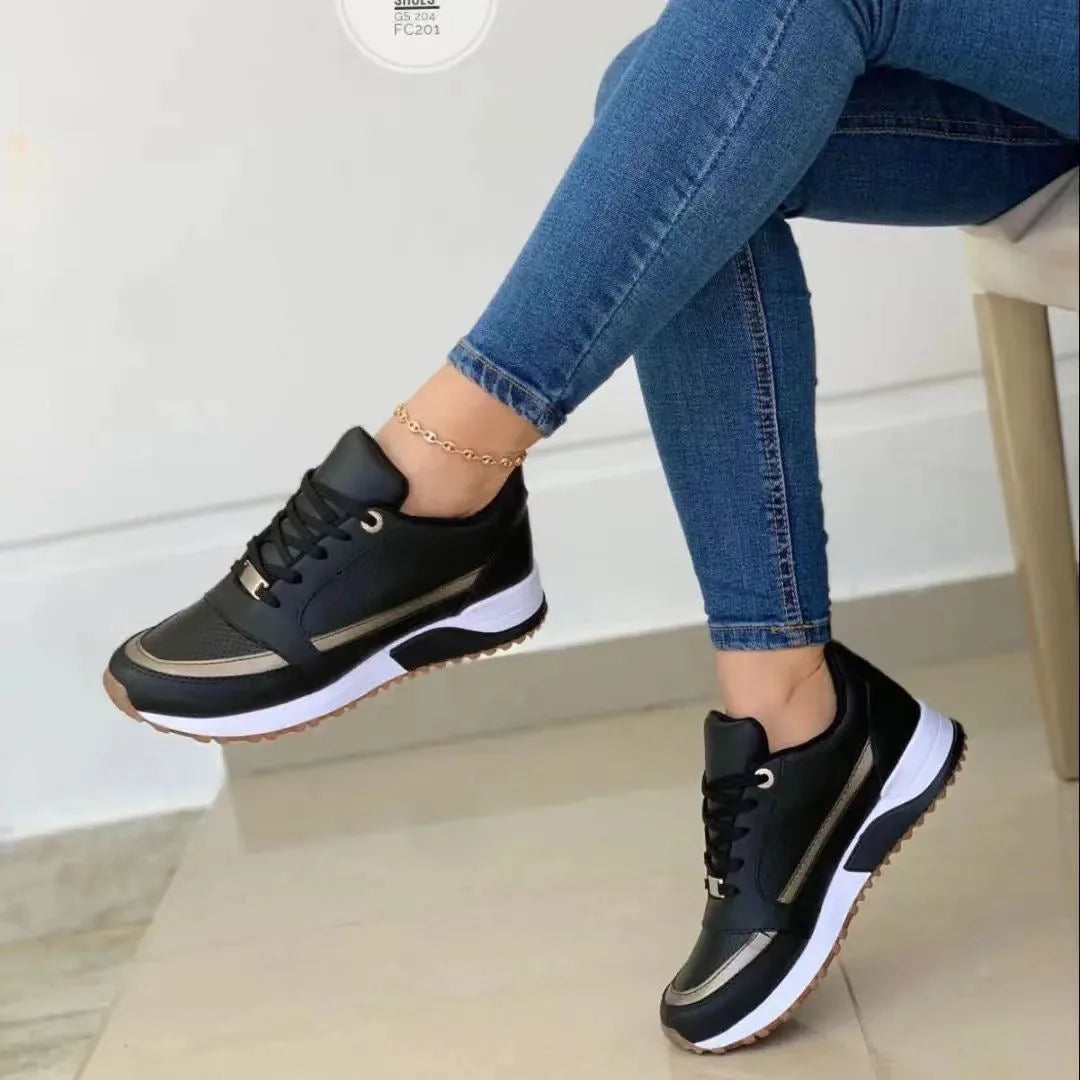 Black trainers with bronze side panel and white chunky sole, shown on a seated model with jeans and a gold anklet