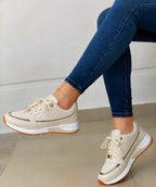 Beige perforated trainer with metallic gold trim along the side panel and white cushioned sole, shown on a seated model's foot.