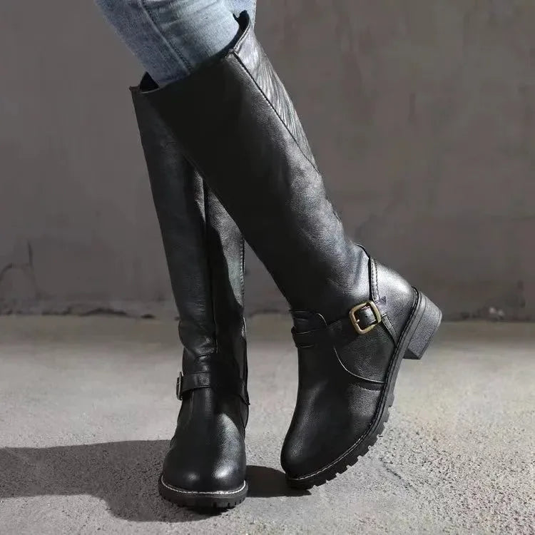 Side view of black knee-high leather boot with buckle strap detail and low block heel, worn with jeans