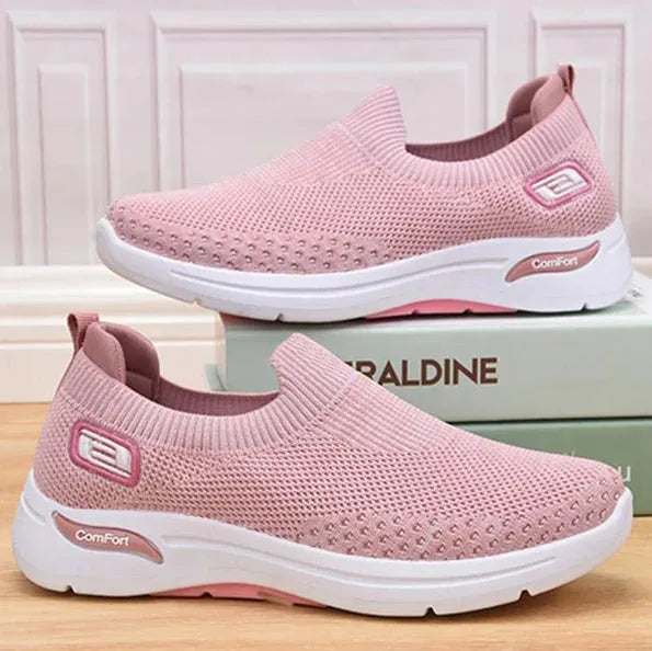 Pair of pink knit slip-on trainers displayed on top of stacked gift boxes against a pastel backdrop