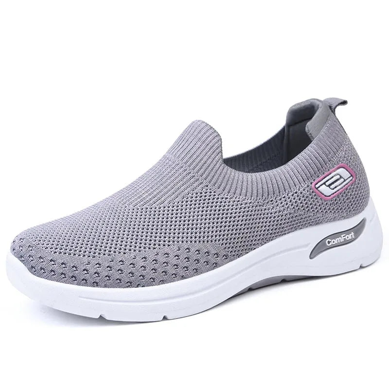 Side angle of a grey knit slip-on trainer with pink accent badge and white cushioned sole