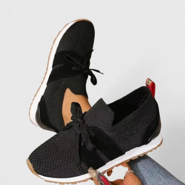 Black knit slip-on sneakers with bow detail and white sole, crossed over each other showing side profile