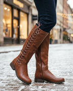 Side view of tan brown lace-up knee-high boot worn with navy jeans on a snowy cobbled street