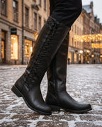 Black lace-up knee-high boot with low block heel shown against snowy cobblestones at dusk