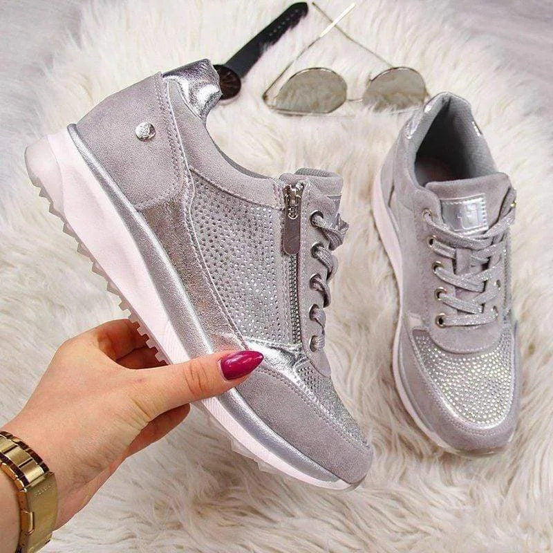 Pair of grey trainers with silver panels and diamante toe resting on a faux fur rug next to a watch and sunglasses