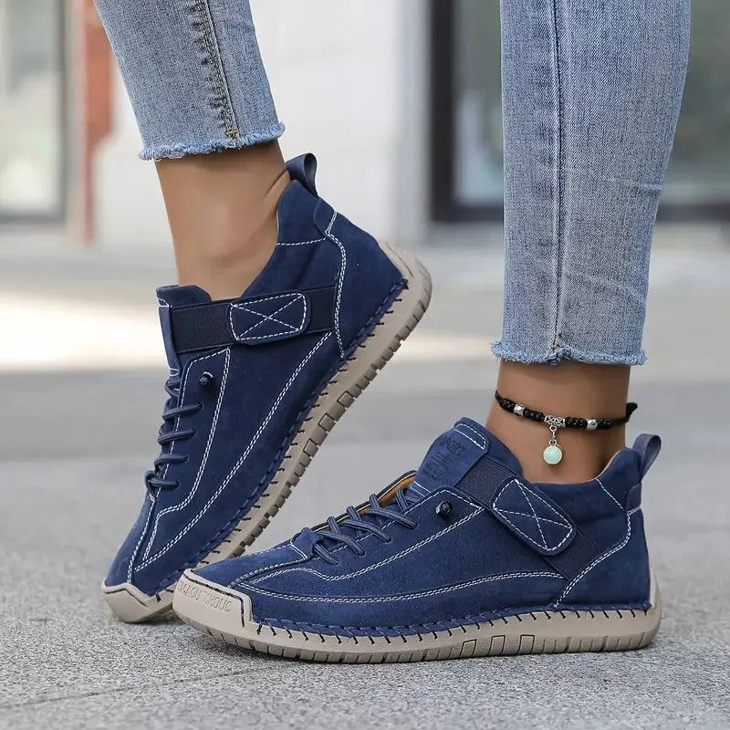 Navy blue suede trainer with tan sole and elastic strap, shown from a low side angle on pavement