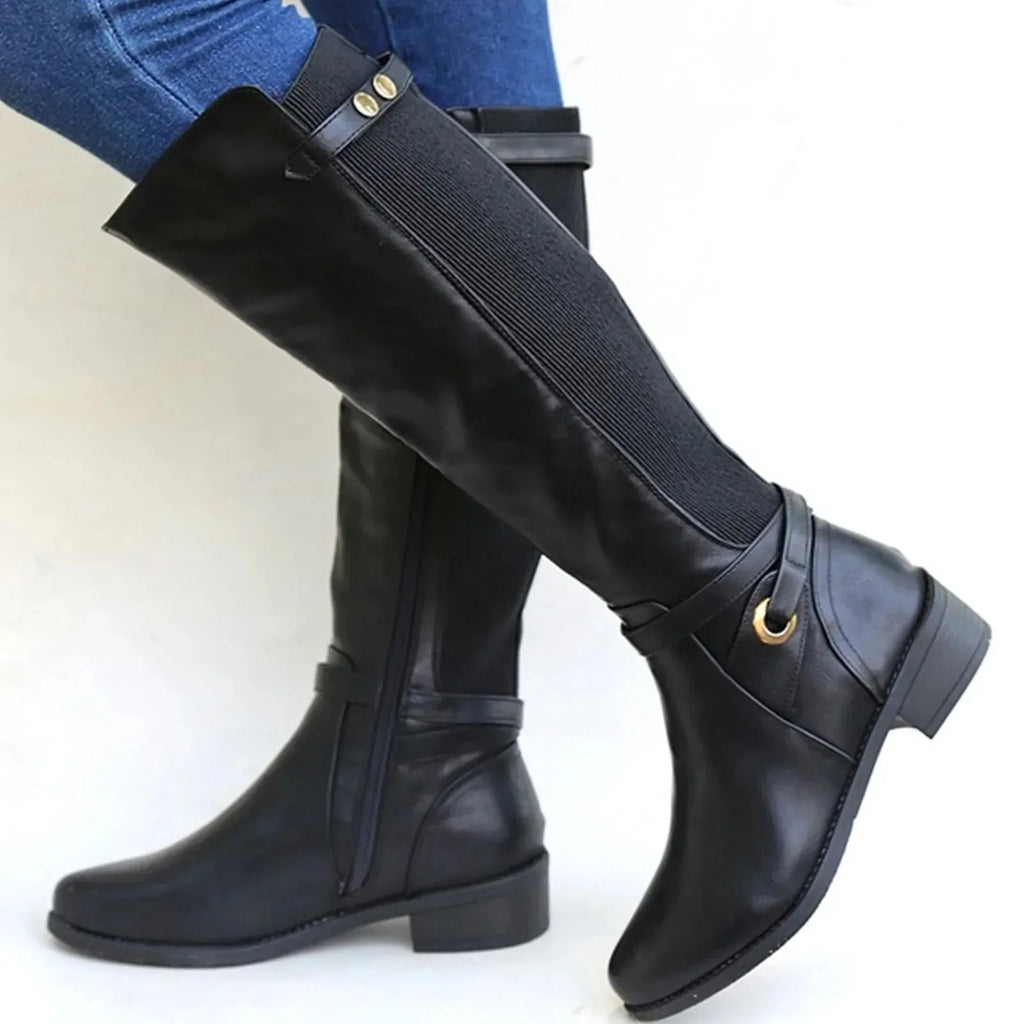 Side view of a black knee-high leather boot with elastic gusset panel and buckle detail, worn with jeans