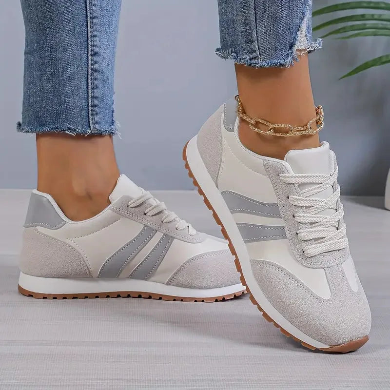 Side view of grey and white lace-up trainers with silver stripe panels, worn with cropped jeans and a gold anklet.
